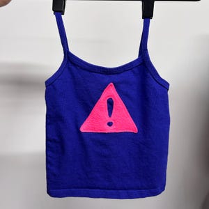 Puede incluir: Un top azul de tirantes con un triángulo rosa que contiene un signo de exclamación. El top tiene tirantes finos.