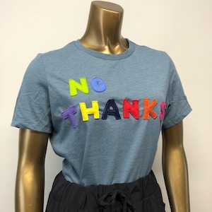 Puede incluir: Una camiseta azul con las palabras "NO THANKS" en letras de fieltro de colores arcoíris.