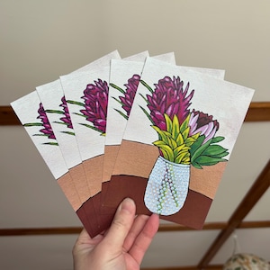 Puede incluir: Cinco tarjetas blancas con una pintura de flores rosas y amarillas en un jarrón blanco con superficie texturizada. El jarrón está sobre una mesa marrón.