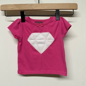 Puede incluir: Una camiseta rosa de manga corta con una aplicación en forma de diamante blanco en la parte delantera.