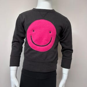 Puede incluir: Una sudadera negra con una gran cara sonriente rosa en la parte delantera. La cara sonriente está delineada en negro.