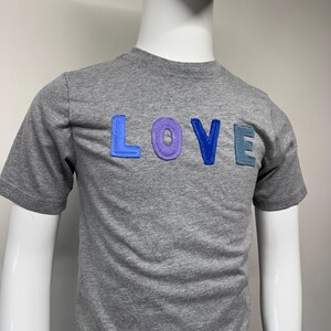 Puede incluir: Una camiseta gris con la palabra "LOVE" aplicada en el pecho. Las letras son en tonos de azul y morado. La camiseta se muestra en un maniquí.