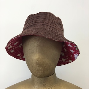 Puede incluir: Un sombrero de cubo de pana marrón con un forro floral rojo y blanco. El sombrero es reversible y se puede usar con cualquiera de los lados hacia afuera.