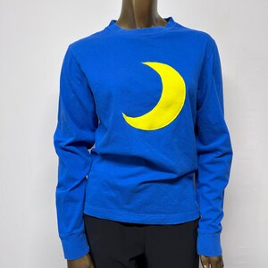 Puede incluir: Una camiseta azul de manga larga con un gráfico de luna creciente amarilla.