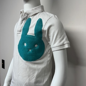 Puede incluir: Camiseta polo blanca de manga corta con un aplique de conejo azul turquesa en la parte delantera.