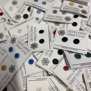 Puede incluir: Una colección de pendientes de botón reciclados en tarjetas blancas. Los pendientes vienen en varios colores, incluyendo negro, azul, rojo y beige. Cada tarjeta está etiquetada como "Criddlebee Creations" e incluye el texto "Upcycled Button Earrings with Stainless Steel Posts & Backs."