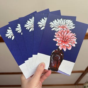 Puede incluir: Una mano sostiene una pila de tarjetas de felicitación con un diseño floral. Cada tarjeta presenta un fondo azul oscuro con una ilustración de flores blancas y rosas en un jarrón marrón. Las tarjetas tienen un borde blanco.