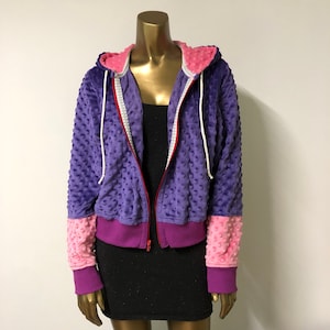 Puede incluir: Sudadera con capucha de color morado, rosa y rojo con tejido texturizado. La sudadera tiene cierre de cremallera y capucha con cordón.