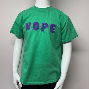 Puede incluir: Camiseta verde de manga corta con la palabra "NOPE" en letras hinchadas de color morado.