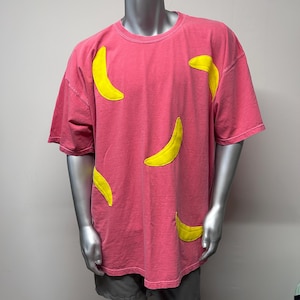 Puede incluir: Una camiseta rosa con aplicaciones de plátanos amarillos. La camiseta tiene cuello redondo y mangas cortas.