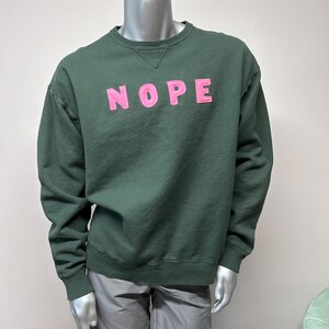 Puede incluir: Una sudadera verde oscuro con un gráfico rosa "NOPE" en la parte delantera.