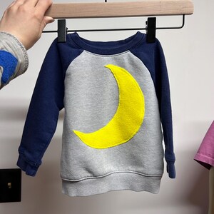 Puede incluir: Una sudadera gris y azul marino con una aplicación de media luna amarilla.