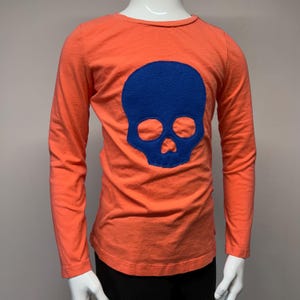 Puede incluir: Una camiseta naranja de manga larga con un diseño de calavera de fieltro azul en la parte delantera.