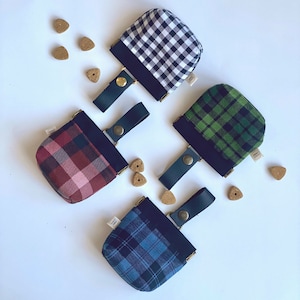 May include: Four dog treat pouches in a variety of plaid patterns: black and white, green, burgundy, and blue. Each pouch has a dark blue trim, a green leather handle, and a gold snap closure. Dog treats are scattered around the pouches.