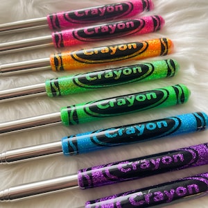 May include: A collection of colorful, glitter-covered pens designed to resemble crayons. Each pen features a different color, including pink, orange, green, blue, and purple, with the word "Crayon" printed on the barrel in a contrasting color. The pens have silver metal shafts.