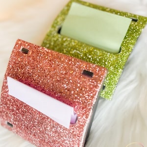 May include: Two glitter-covered sticky note holders, one in rose gold and the other in lime green. Each holder has a rectangular opening to dispense sticky notes. The holders are on a white, fluffy surface.