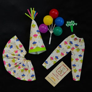 May include: A birthday party outfit for a doll, featuring a white skirt and pants with cupcake designs, a green and white cupcake hat, and a set of colorful balloons with a "Happy Birthday" topper.