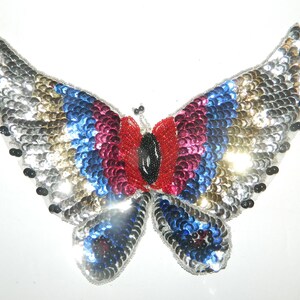 May include: A silver, gold, blue, and red sequined butterfly applique. The butterfly has a black beaded body and black beaded antennae.