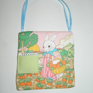 May include: A square fabric handbag with a light blue ribbon handle. The bag features a colorful illustration of a white rabbit in pink overalls holding a basket of carrots in a field. A green pocket is sewn on the front.