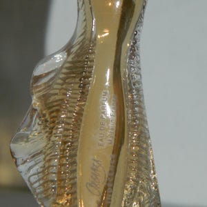 May include: A clear glass perfume bottle with a gold-colored stopper and a gold-colored interior. The bottle has a unique, sculpted shape and the text "Organza Eau de Parfum Made in France" is visible.