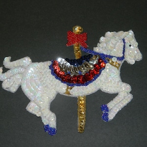 May include: A white sequined carousel horse with a red, white, and blue saddle and a gold pole. The horse is standing on a black background.