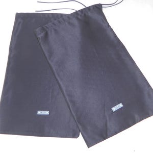May include: Two dark gray fabric dust bags with drawstring closures. Each bag has a small rectangular label with the brand name "PRADA" in white lettering. The bags are likely for protecting luxury items.