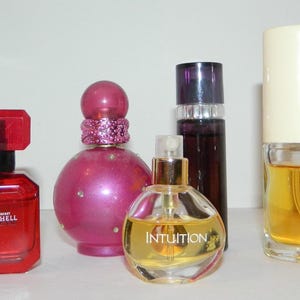 May include: An assortment of perfume bottles in various shapes and sizes. The bottles are red, pink, purple, and clear, with gold and black accents. One bottle is labeled "Bombshell" and another "Intuition".
