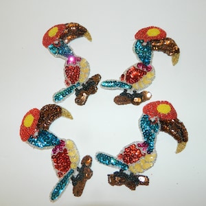 May include: Four colorful toucan bird appliques made of sequins. The toucans are in various shades of blue, red, yellow, and brown. Each toucan has a yellow beak and a red head.