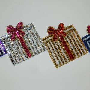 May include: Four sequined gift box patches in different colors. The boxes are decorated with a bow and a stripe pattern. The colors are purple, silver, gold, and blue.