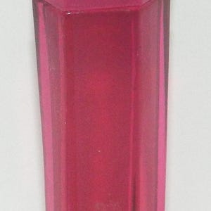 May include: A tall, slender, translucent magenta perfume bottle with a faceted, hexagonal glass body and a matching angled cap. A white infinity symbol logo is printed near the base of the bottle, which features clean, vertical lines and a vibrant, deep pink hue.