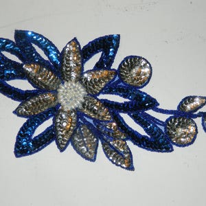 May include: A blue and silver sequined flower applique with a pearl center. The applique is perfect for adding a touch of sparkle to clothing, bags, or home decor.