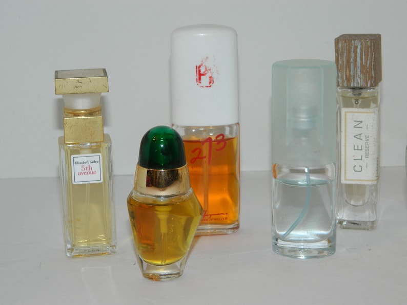 Puede incluir: Una colecci&oacute;n de frascos de perfume de varias formas y tama&ntilde;os. Los frascos contienen l&iacute;quidos de diferentes colores, incluyendo amarillo y naranja. Un frasco tiene una tapa dorada y una etiqueta que dice "5th avenue". Otro frasco tiene una tapa verde.