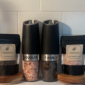 May include: Two black electric salt and pepper grinders with clear glass containers filled with pink Himalayan salt and black peppercorns. The grinders are labeled "DOUGLAS" and "FAMILY". The grinders are on a wooden surface with small bags of salt and pepper labeled "STARTER PACK" with the type of salt and pepper listed.