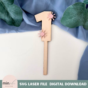 May include: A wooden number one with two pink sun-shaped embellishments. The number is on a white background.