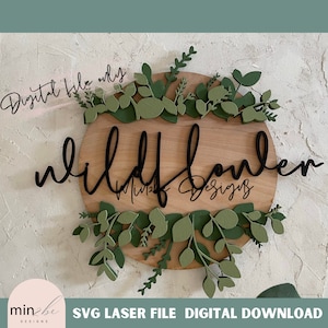 Könnte beinhalten: Ein Holzschild mit dem Text "wildflower" in schwarzer Schrift. Das Schild ist von einem Kranz aus grünen Blättern umgeben.