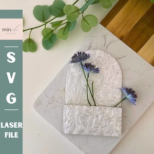 May include: White decorative wall pocket with a rounded top and a rectangular opening. The pocket is made of a white material with a marbled pattern. Two purple flowers are in the opening.