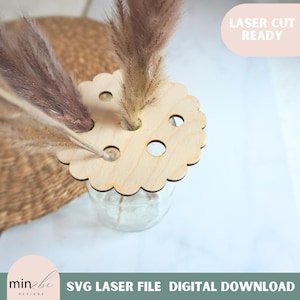 May include: A laser cut wooden flower vase topper with multiple holes for stems. The topper is in the shape of a circle with scalloped edges. The topper is on top of a clear glass vase with dried pampas grass.