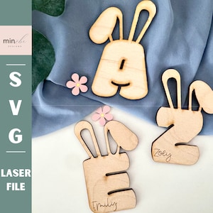 May include: Three wooden letters, A, E, and Z, shaped like bunnies. The letters are painted in a natural wood color and have the names "Emily", "Zoey" and "A" written on them. The letters are on a blue fabric background.