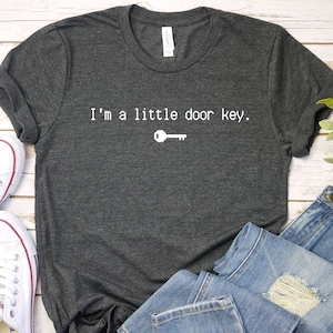 Könnte beinhalten: Ein dunkelgraues T-Shirt mit dem Text "I'm a little door key." in weißer Schrift und einem kleinen weißen Schlüsselsymbol.