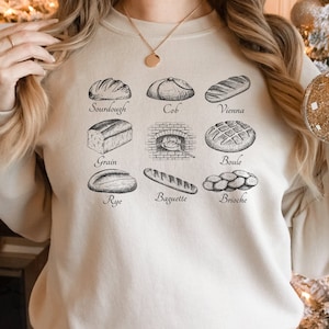 May include: A beige sweatshirt with a graphic of different types of bread, including sourdough, cob, vienna, grain, rye, baguette, boule, and brioche.