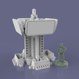 May include: A 3D-printed model of a futuristic structure with a rectangular screen and a small figure of a soldier. The structure is light gray with detailed mechanical elements. The soldier figure is green and stands on a circular base.