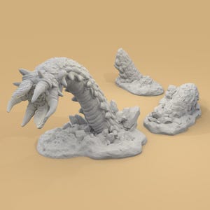 May include: A gray, 3D-printed model of a sandworm emerging from a rocky terrain, with a detailed head and segmented body. Two additional rock formations are also present, suggesting a fantasy or tabletop gaming theme.