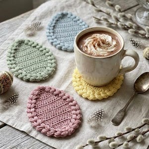 Könnte beinhalten: Eine Tasse heiße Schokolade mit Schlagsahne auf einem gelben gehäkelten Untersetzer. Vier pastellfarbene, eiförmige Untersetzer umgeben die Tasse. Dekorative Ostereier und Federn liegen ebenfalls auf dem Tisch.