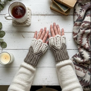 May include: A pair of cream and brown fingerless gloves with a textured crochet design. The gloves are paired with a cream sweater, a cup of tea, a crochet coaster, a candle, a stack of books, and a patterned blanket.