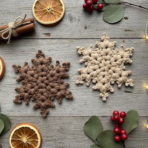 Op de afbeelding: Twee gehaakte sneeuwvlokonderzetters, één bruin en één beige, liggen op een verweerd houten oppervlak. Gedroogde sinaasappelschijfjes, kaneelstokjes en takjes groen met rode bessen voegen een feestelijke toets toe. Er zijn ook lichtjes te zien.