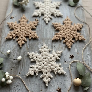 Op de afbeelding: Vijf handgemaakte gehaakte sneeuwvlokken in tinten wit en bruin zijn gerangschikt op een verweerd grijze houten ondergrond. Decoratieve elementen zijn takjes groen, witte bessen en kleine stervormige ornamenten.