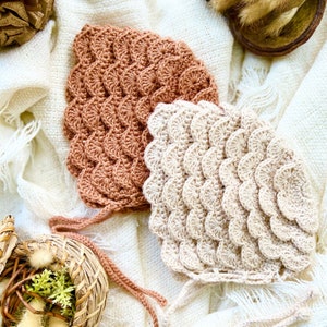 May include: Two crocheted baby bonnets in a beige and terracotta color scheme. The bonnets are made with a textured, scalloped stitch.