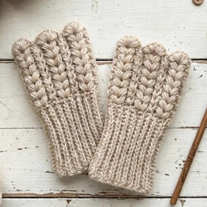 May include: A pair of beige knitted fingerless gloves with a textured cable knit design. The gloves are displayed with several balls of yarn, wooden buttons, a wooden crochet hook, and a wooden spool on a white wooden surface.