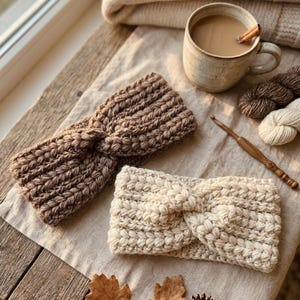 May include: Two handmade, knitted headbands in brown and cream colors are displayed on a wooden surface. A mug of coffee with a cinnamon stick, yarn, and a crochet hook are also visible, suggesting a cozy, handcrafted theme.
