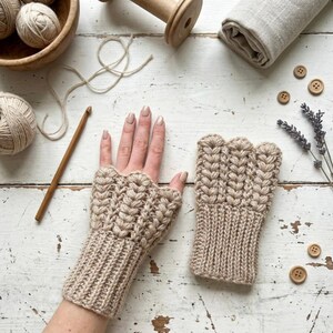 May include: A pair of beige fingerless gloves with a textured, woven design. The gloves are displayed on a white wooden surface, alongside yarn, a crochet hook, wooden buttons, and lavender sprigs. The gloves are a cozy accessory.